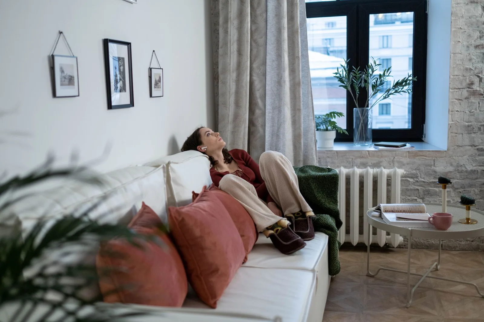 Woman reclining on a white sofa with coral cushions in a bright living room beside a window and radiator