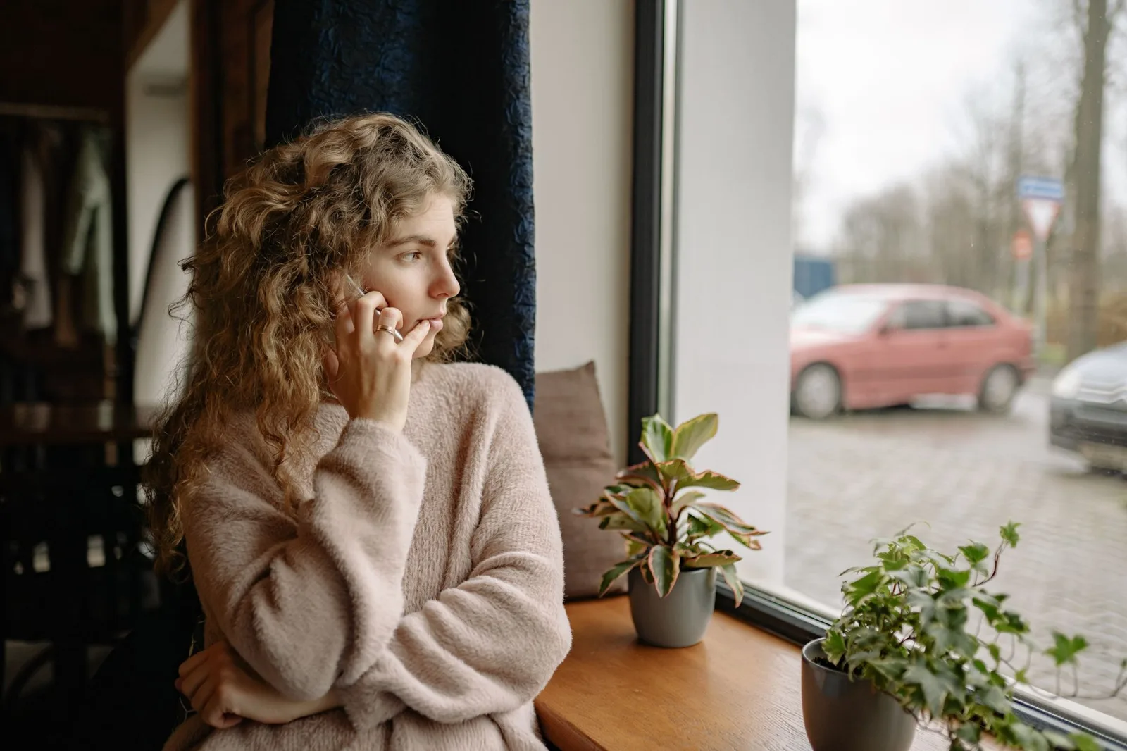 Woman with curly hair in a soft cream jumper sits by a window, hand near her face, looking out at a street