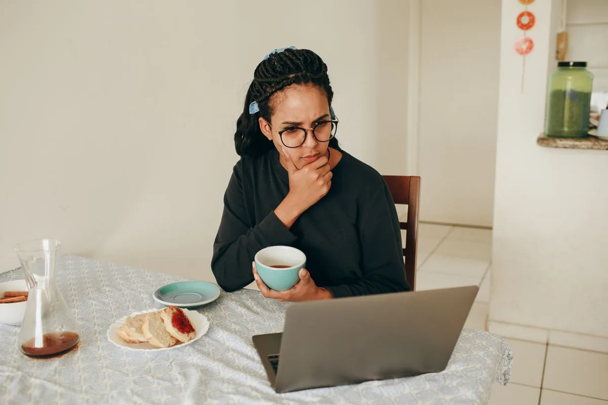 Woman at a kitchen table holding a mug and looking thoughtfully at her laptop